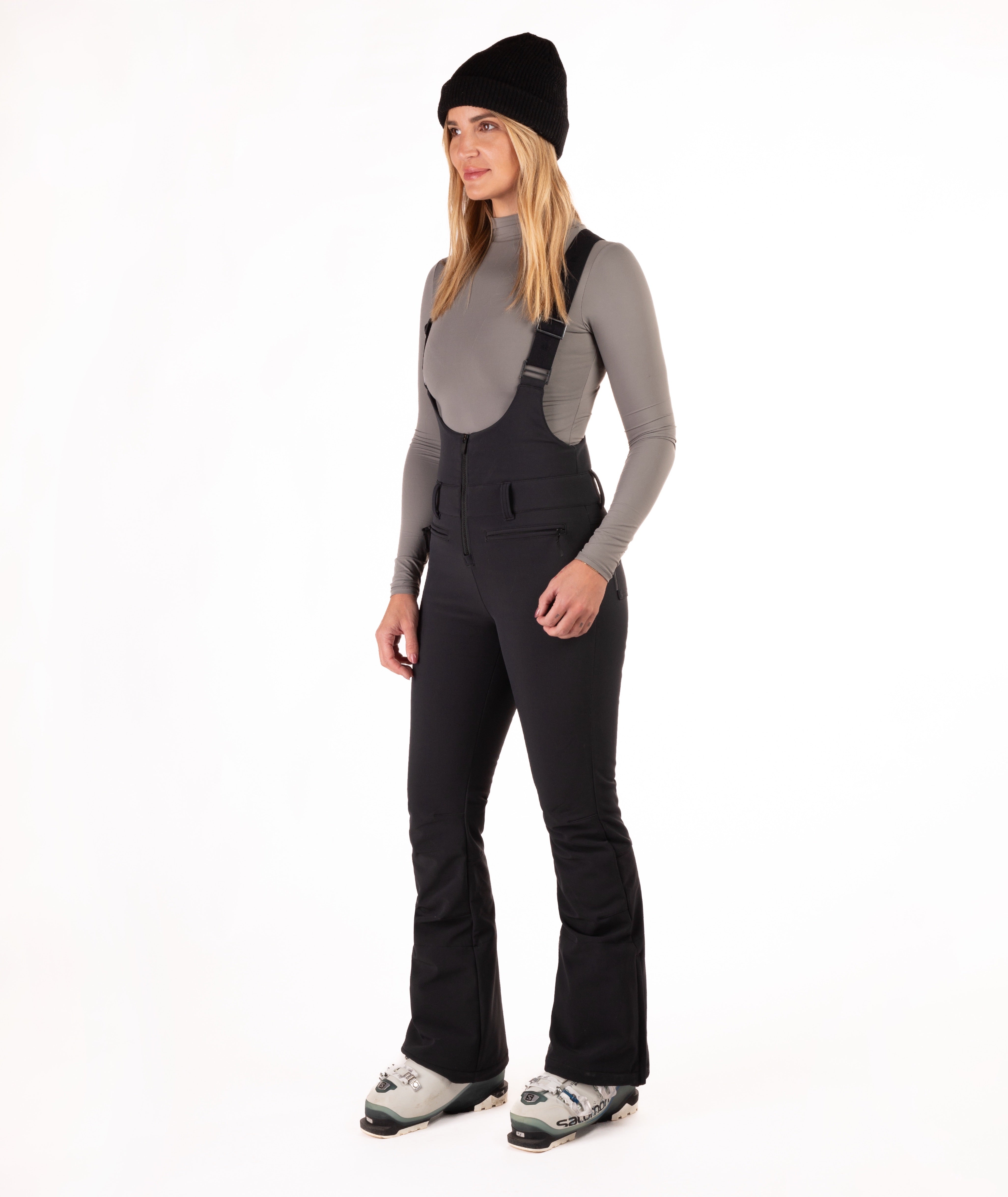 Woman wearing black ski bib overalls with a gray top on a white background