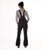 back of a Woman wearing black ski bib overalls with a gray top on a white background