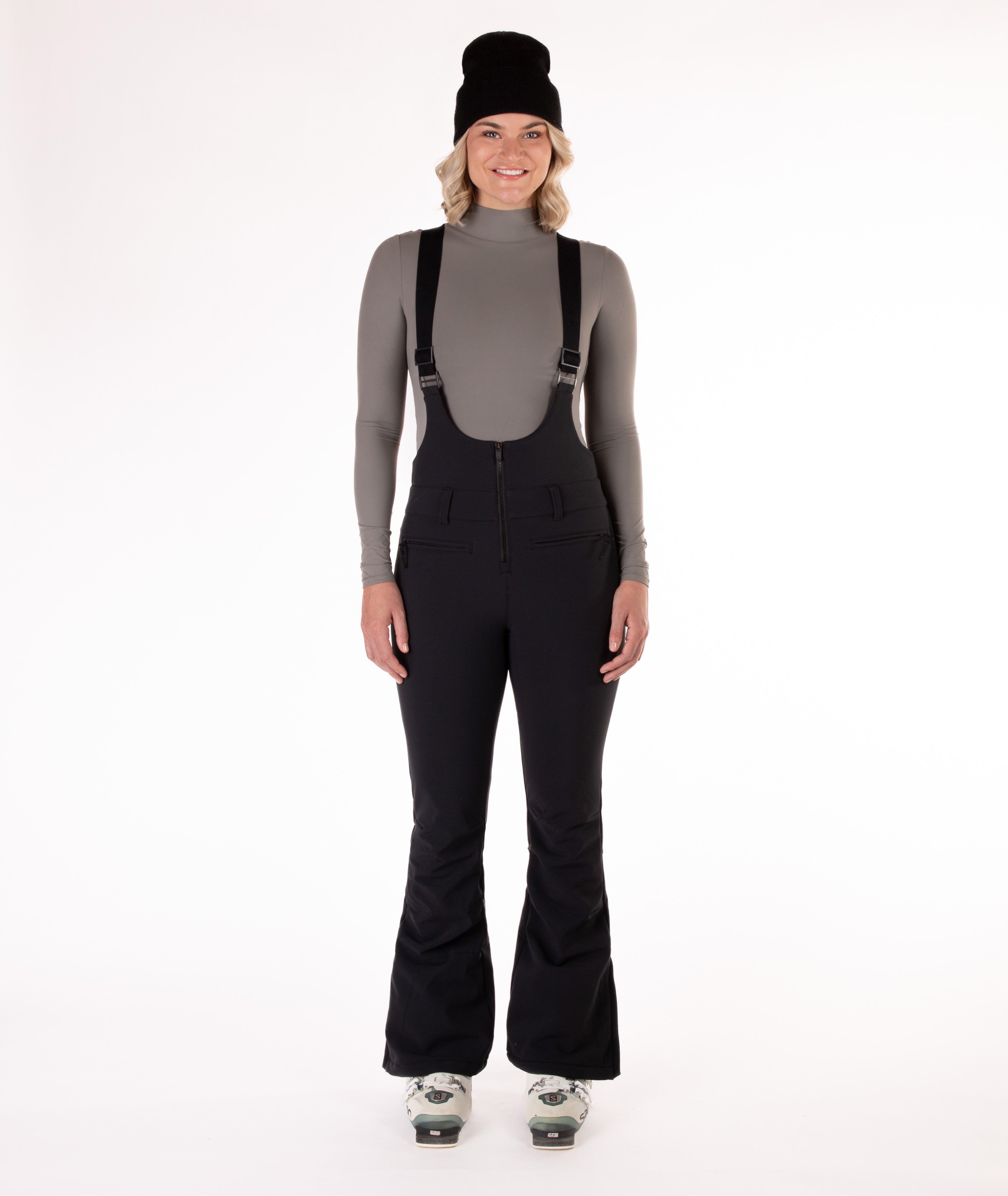 Woman wearing black ski bib overalls with a gray top on a white background