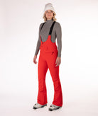Woman wearing red ski bib overalls with a gray top on a white background