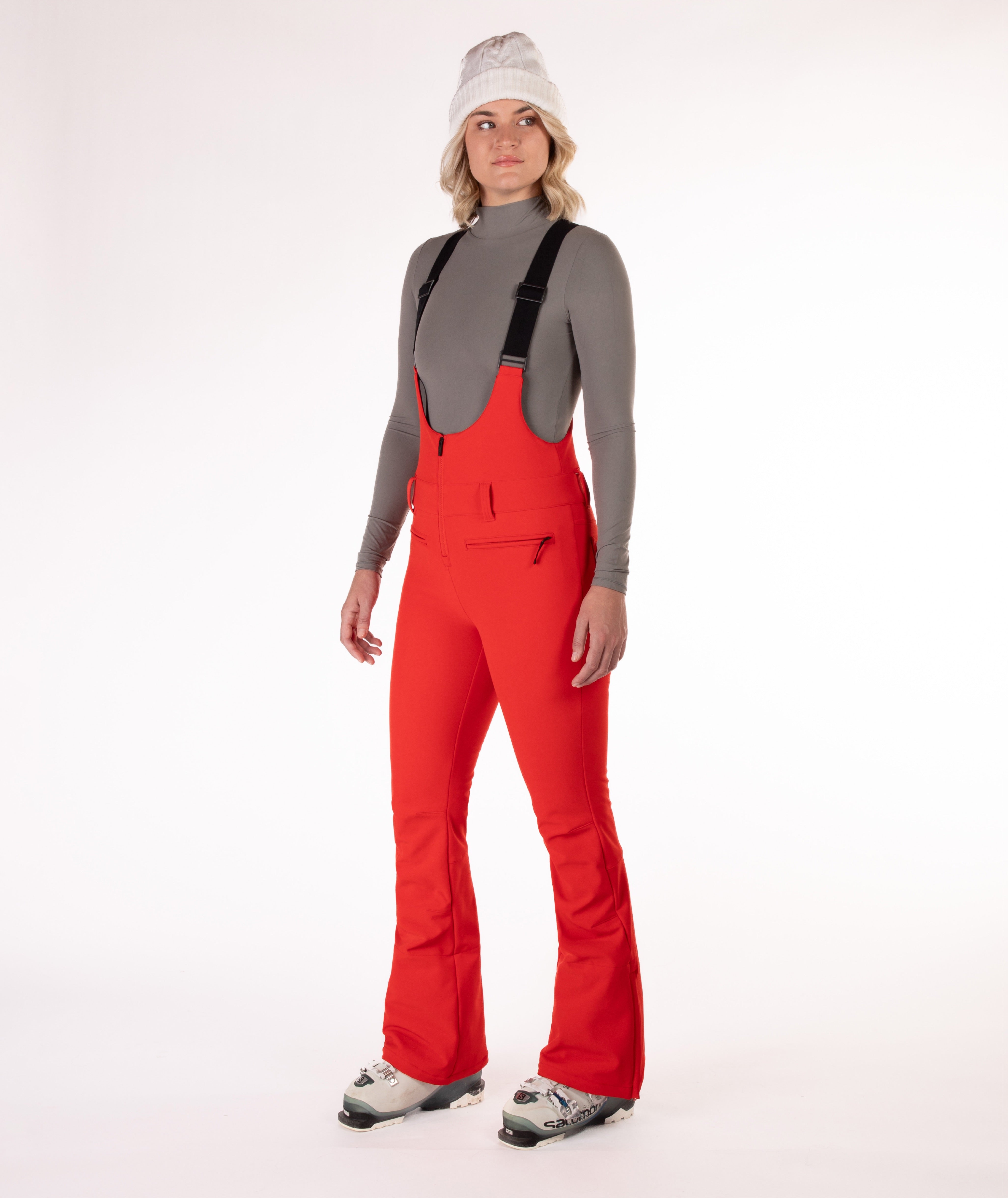 Woman wearing red ski bib overalls with a gray top on a white background