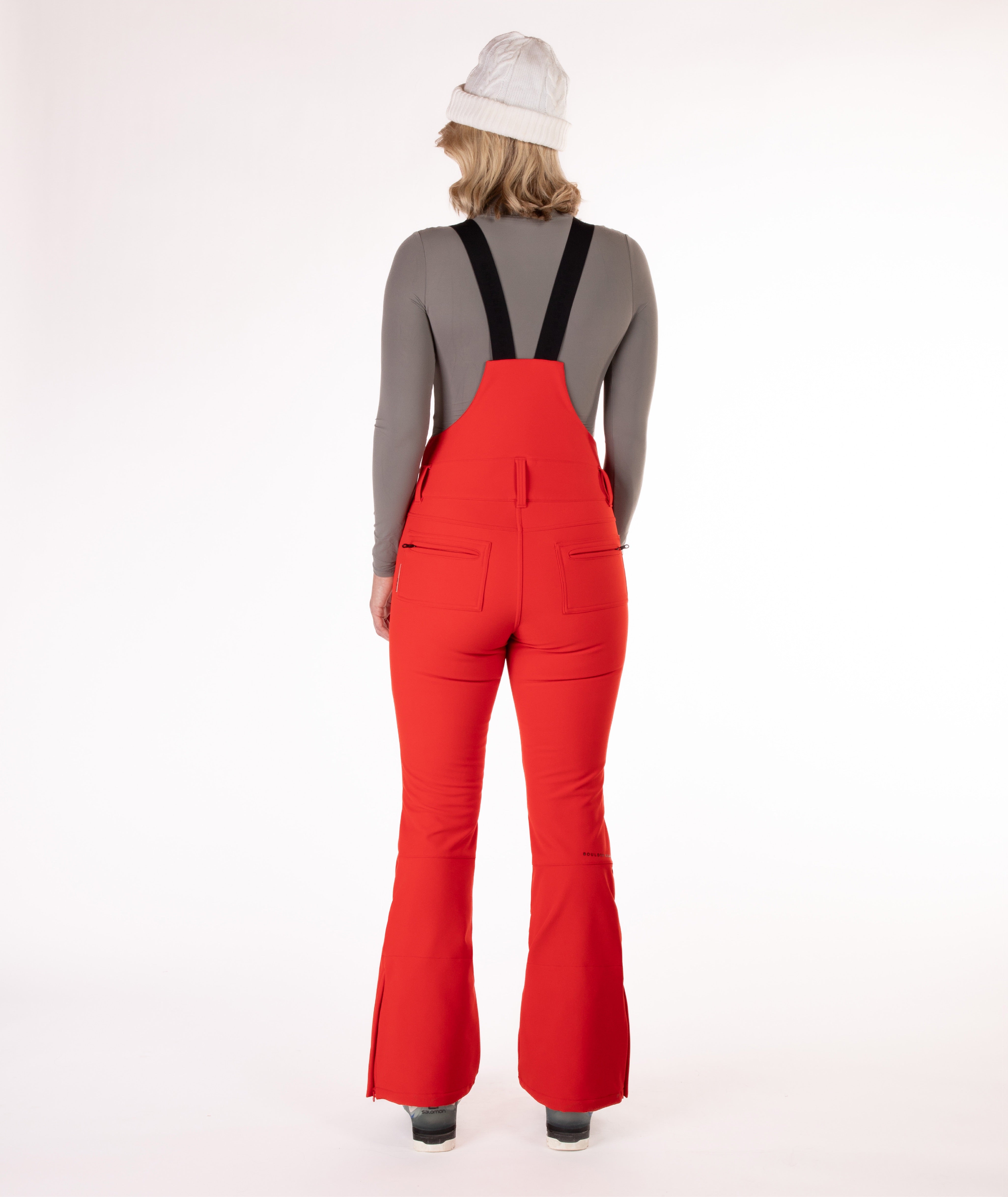 back of a Woman wearing red ski bib overalls with a gray top on a white background