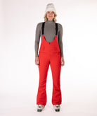 Woman wearing red ski bib overalls with a gray top on a white background
