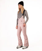 Woman wearing pink ski bib overalls with a gray top on a white background