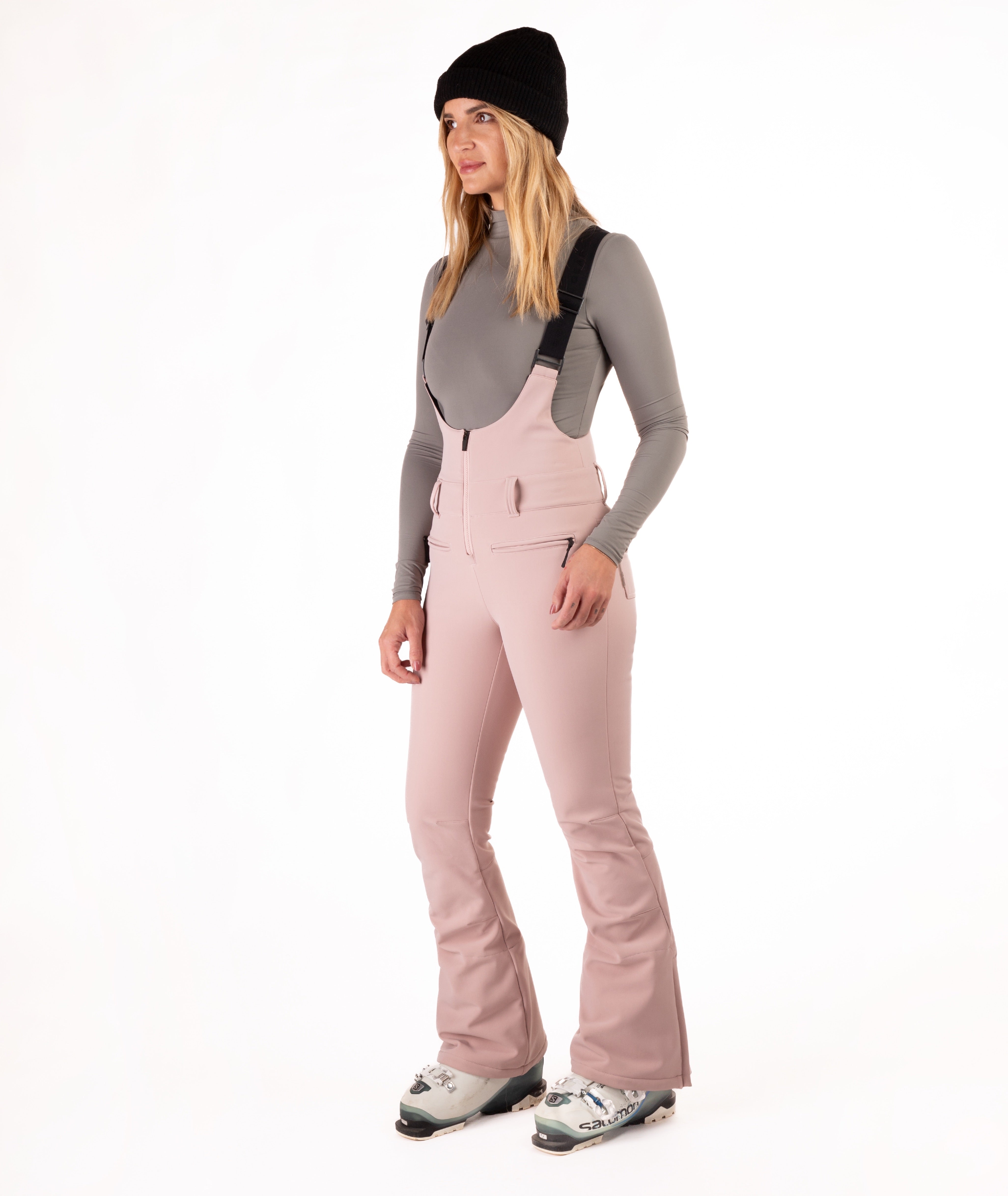 Woman wearing pink ski bib overalls with a gray top on a white background