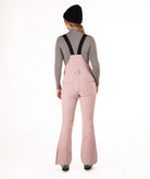 back of a Woman wearing pink ski bib overalls with a gray top on a white background