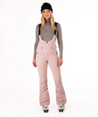 Woman wearing pink ski bib overalls with a gray top on a white background