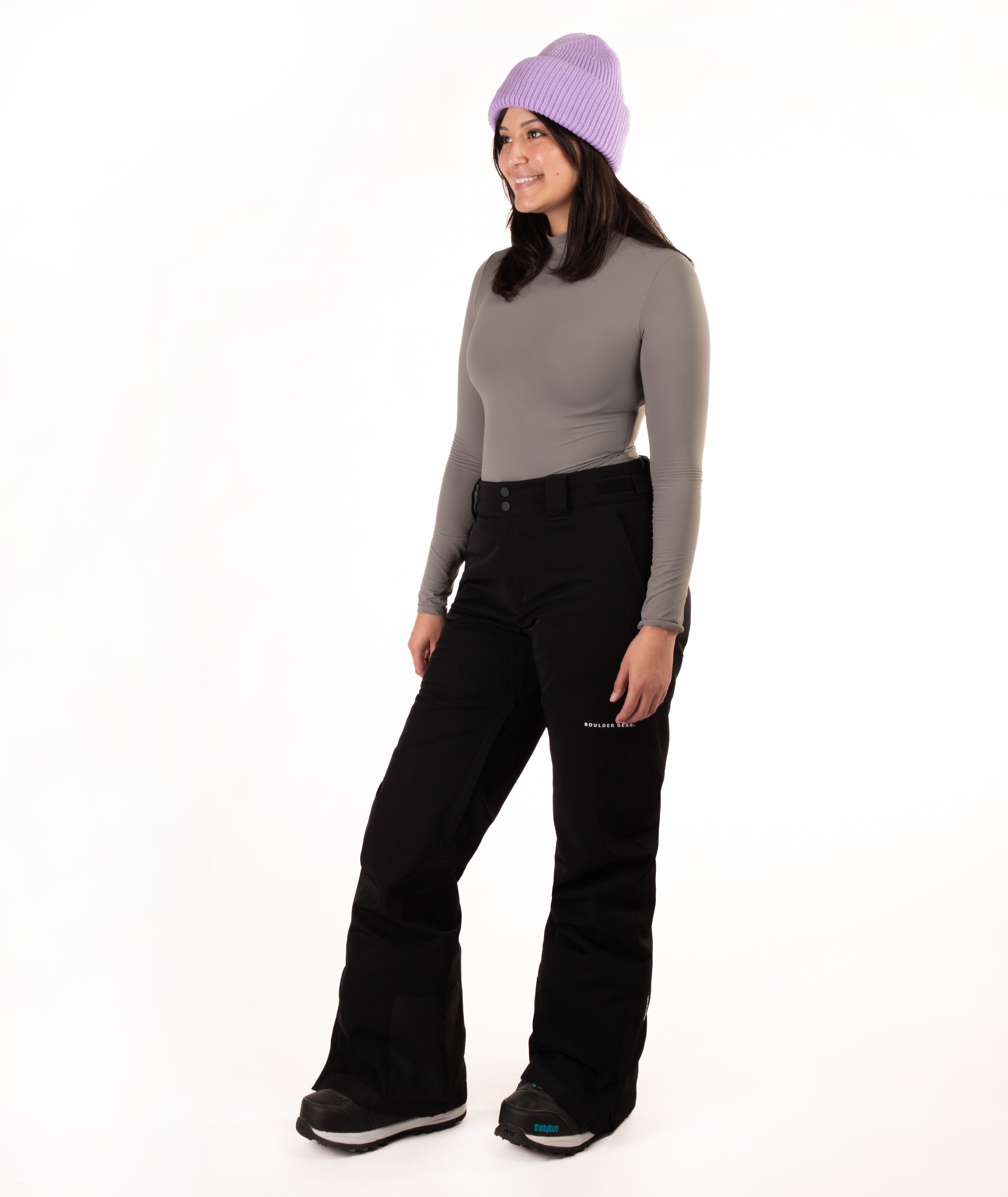 A woman modeling a gray long-sleeve mock neck top, black snow pants, and a light purple knit beanie against a plain white studio background.