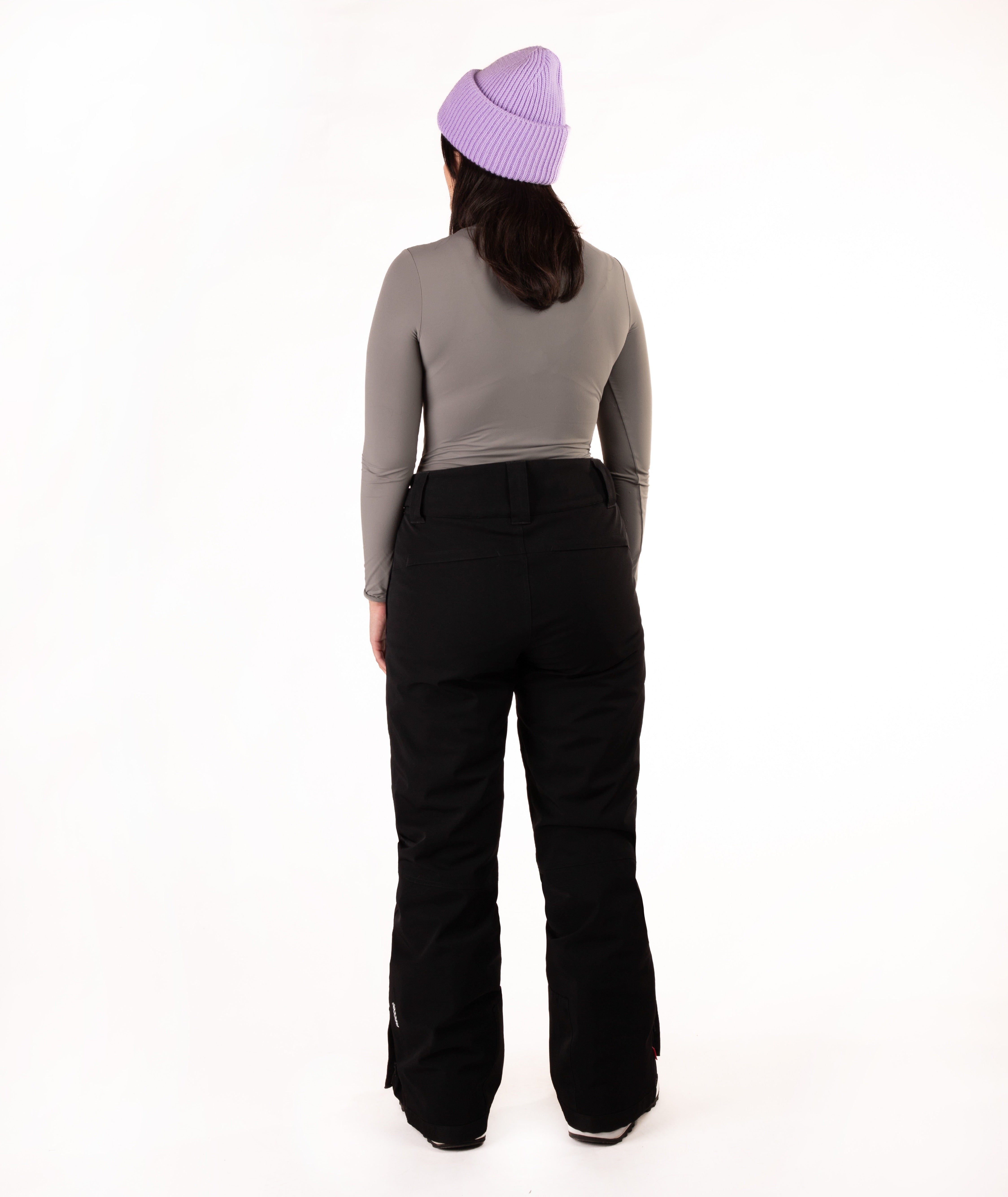 A person shown from the back wearing a light purple knit beanie, a gray long-sleeve top, and black ski pants against a plain white studio background.