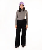 A woman modeling a gray long-sleeve mock neck top, black ski pants, and a lavender knit beanie against a plain white background.