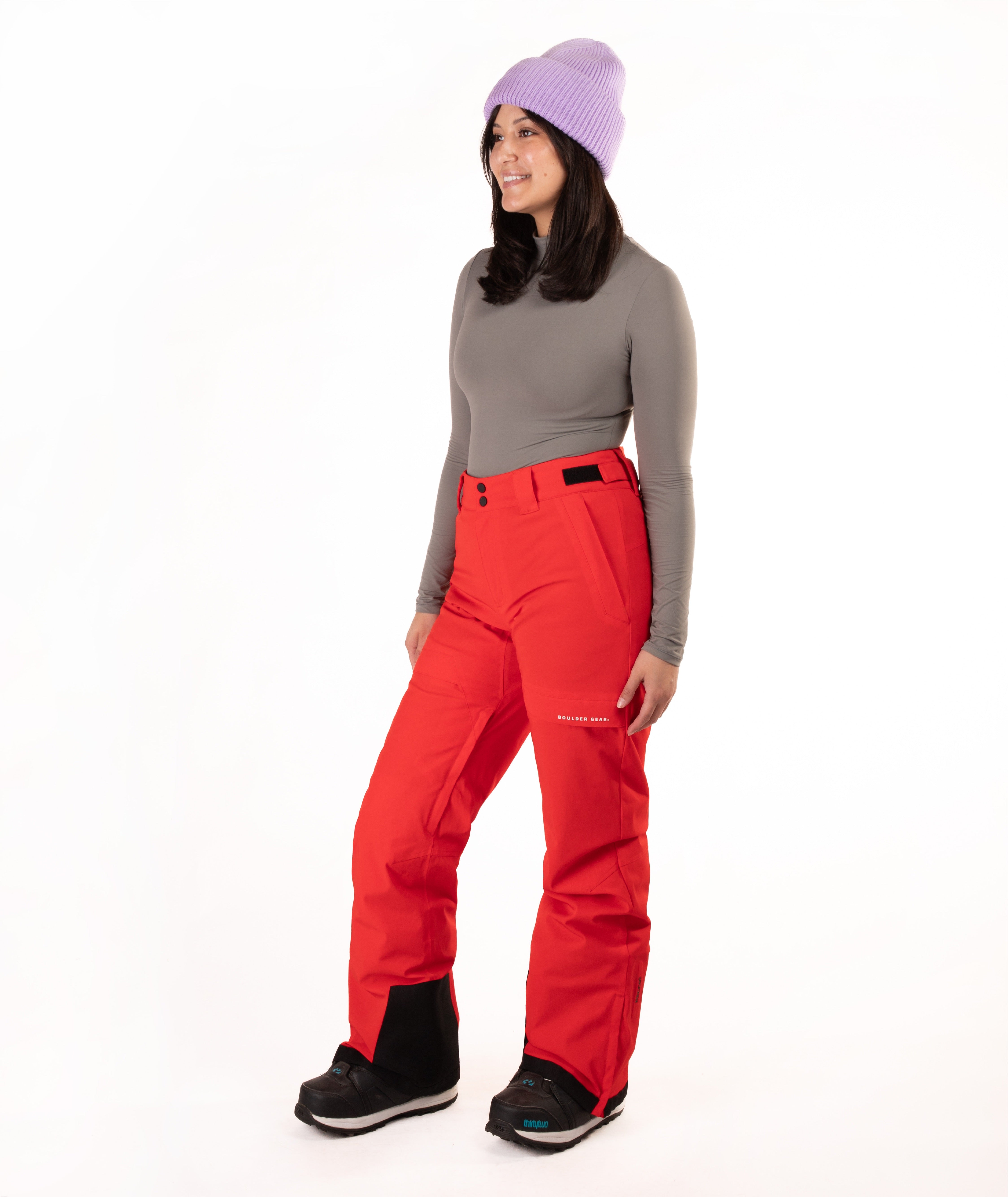 A woman modeling bright red ski pants with a gray long-sleeve top, a light purple knit beanie, and black snow boots, standing against a plain white studio background.