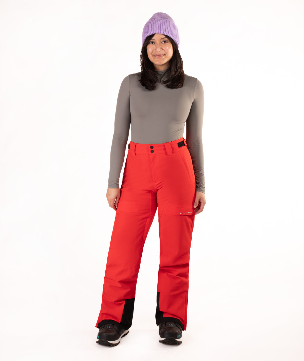 A woman modeling bright red ski pants with a gray long-sleeve top, a purple knit beanie, and black ski boots against a plain white studio background.