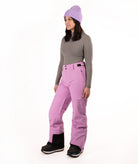 A woman modeling a pair of bright lilac ski pants, paired with a gray long-sleeve top, a lavender knit beanie, and black snow boots, set against a plain white studio background.