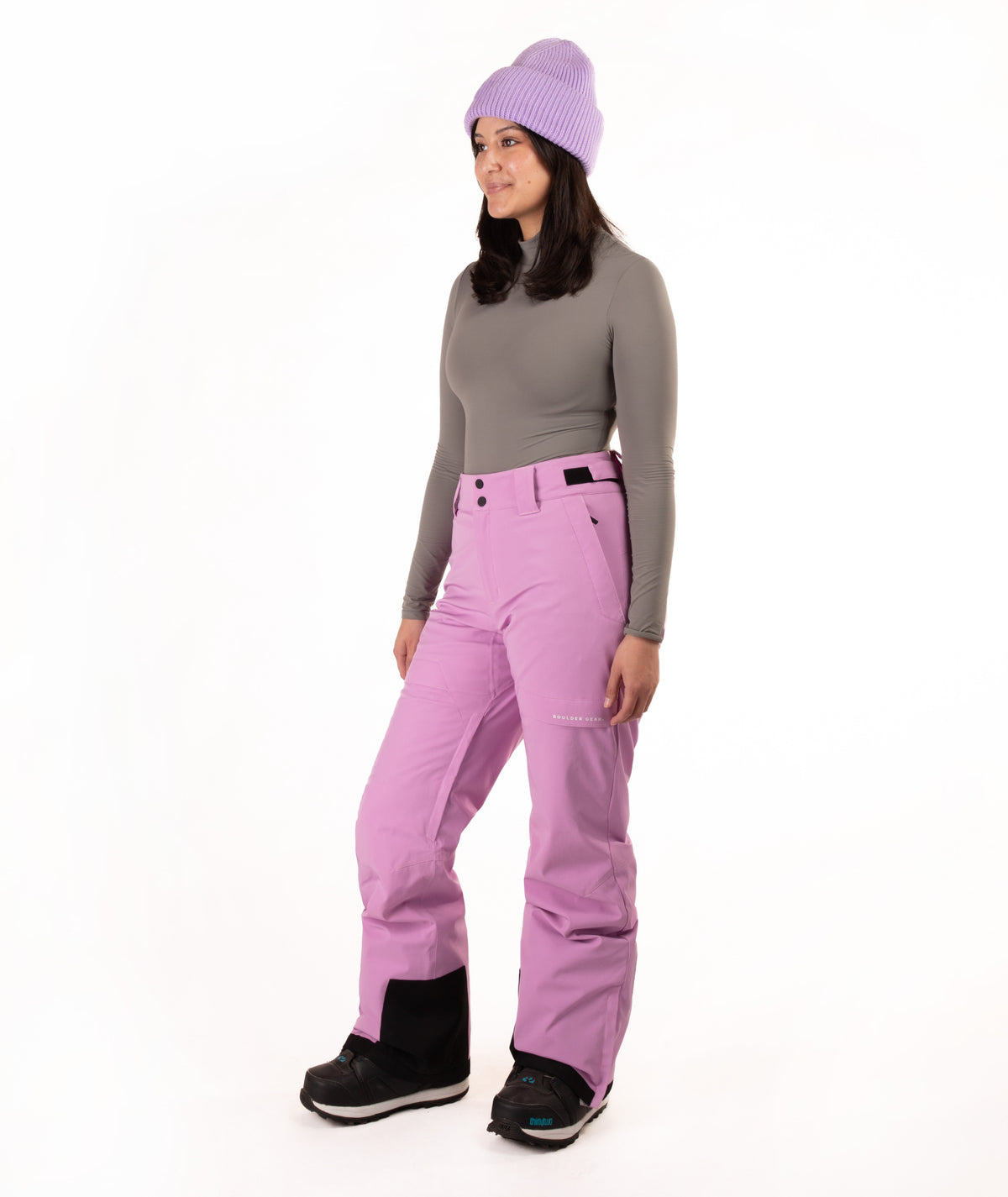 A woman modeling a pair of bright lilac ski pants, paired with a gray long-sleeve top, a lavender knit beanie, and black snow boots, set against a plain white studio background.