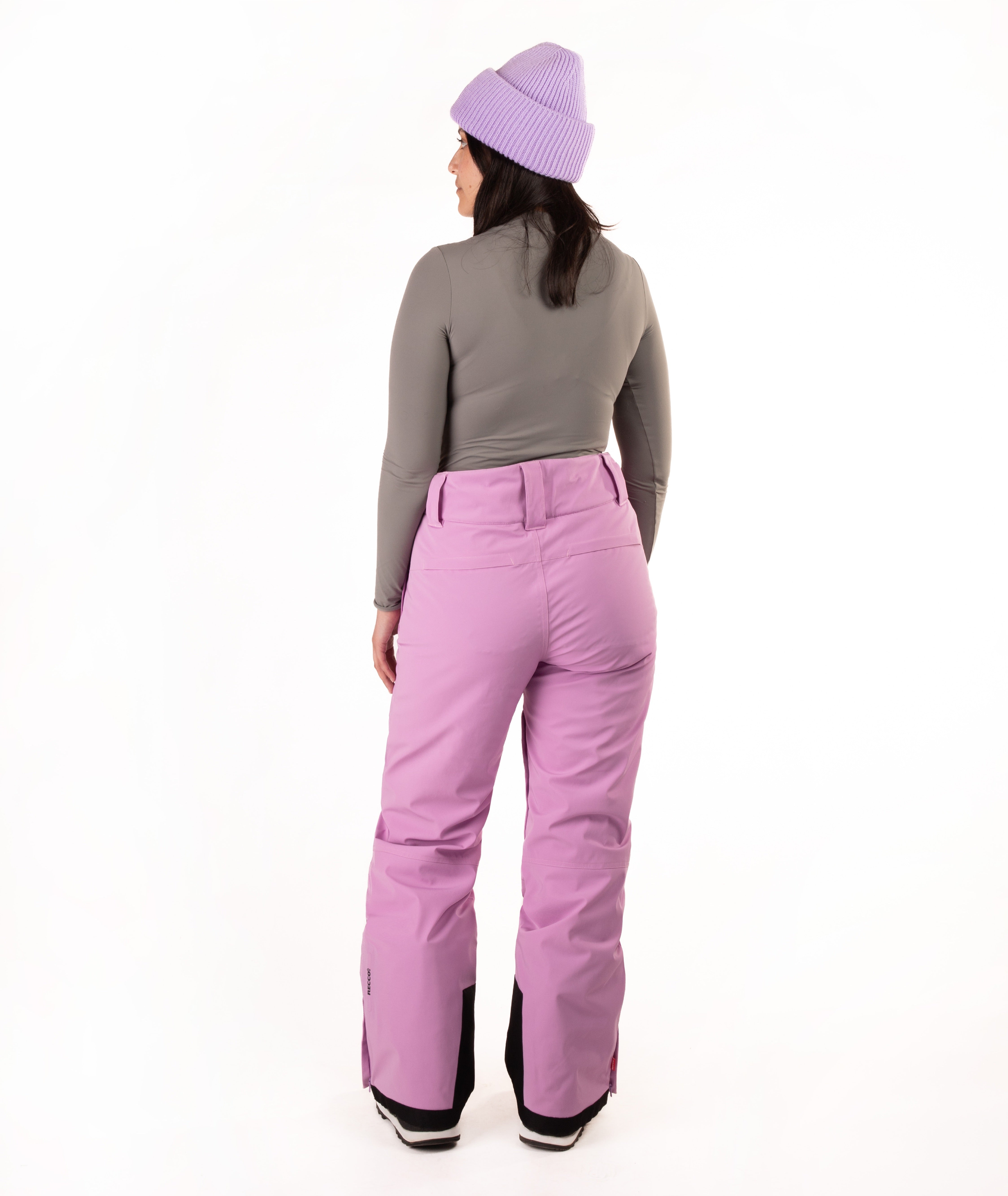 A woman viewed from the back wearing lavender snow pants, a gray long-sleeve top, and a light purple knit beanie against a plain white studio background.