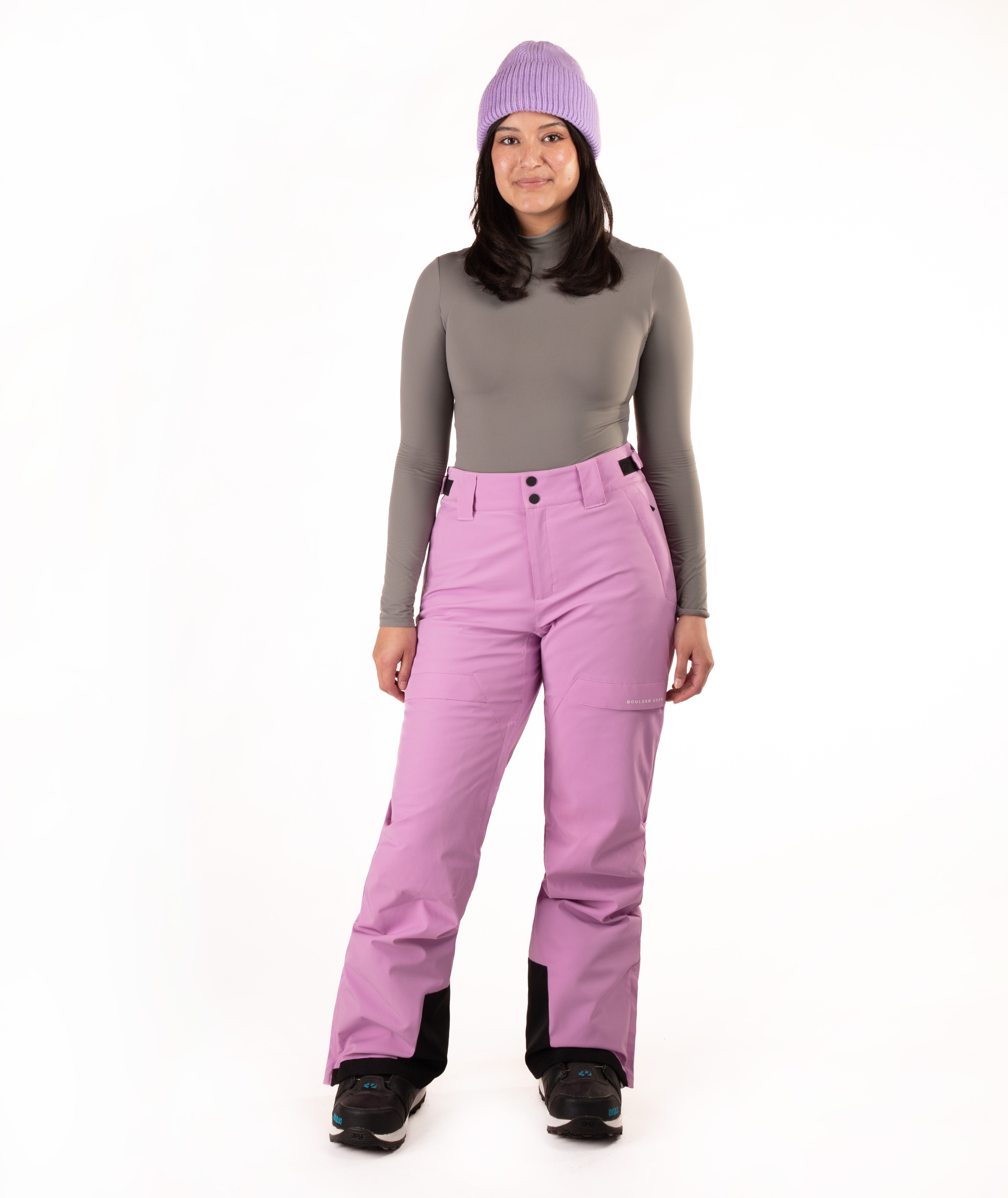A woman modeling a pair of bright lavender-pink ski pants with a matching knit beanie, a gray long-sleeve top, and black snow boots, photographed against a seamless white studio background.