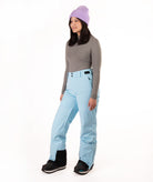 A woman modeling light blue snow pants, a gray long-sleeve top, a purple knit beanie, and black snow boots against a plain white studio background.