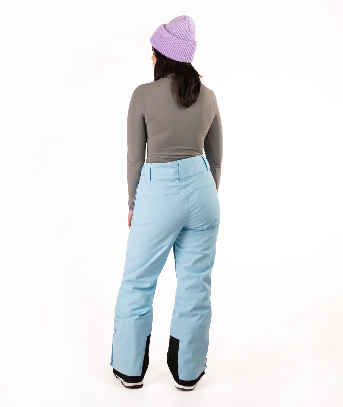 A person shown from the back wearing light blue snow pants, a gray long-sleeve top, and a lavender knit beanie against a plain white studio background.