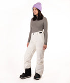 A woman modeling a gray long-sleeve top, off-white snow pants, black snow boots, and a light purple knit beanie against a plain white studio background.