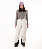A woman modeling a gray long-sleeve top, off-white snow pants with black knee panels, black snowboard boots, and a purple knit beanie against a plain white studio background.