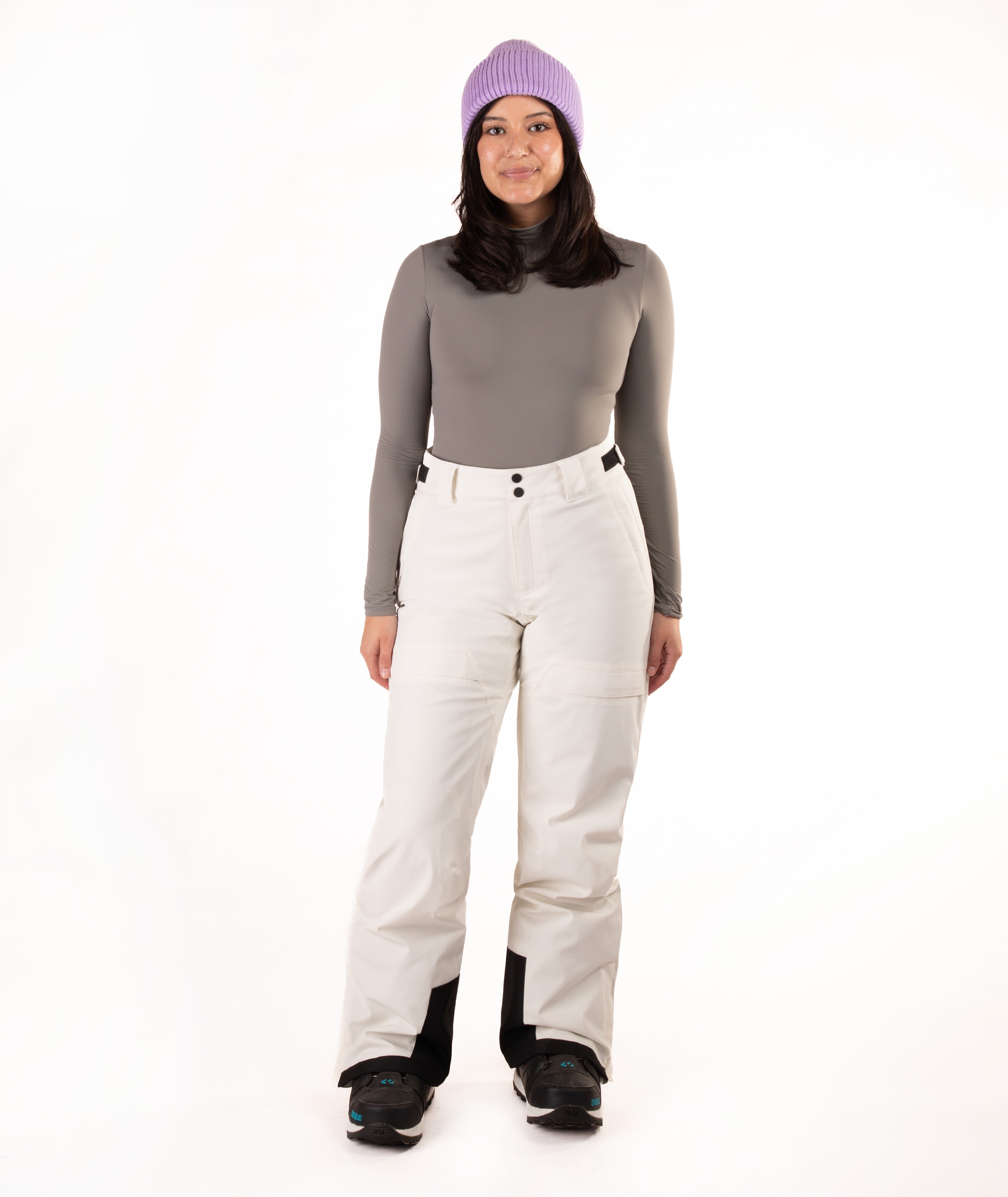 A woman modeling a gray long-sleeve top, off-white snow pants with black knee panels, black snowboard boots, and a purple knit beanie against a plain white studio background.