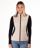 Woman wearing a beige puffer vest over a black long-sleeve shirt on a white background