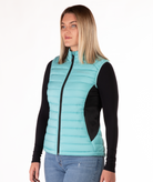 Woman wearing a turquoise puffer vest over a black long-sleeve shirt on a white background