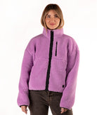 A woman wearing a lavender purple sherpa fleece jacket with a black front zipper and a vertical zip pocket on the chest, paired with dark brown jeans, against a plain white background.