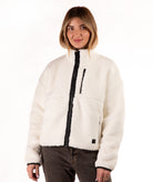 A woman wearing a off-white sherpa fleece zip-up jacket with black trim and a black front zipper, paired with dark brown jeans, against a plain white background.