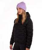 A woman wearing a black hooded puffer jacket and a light purple ribbed knit beanie, photographed against a plain white studio background.