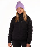 A woman wearing a black puffer jacket and a light purple ribbed knit beanie, standing against a plain white background.