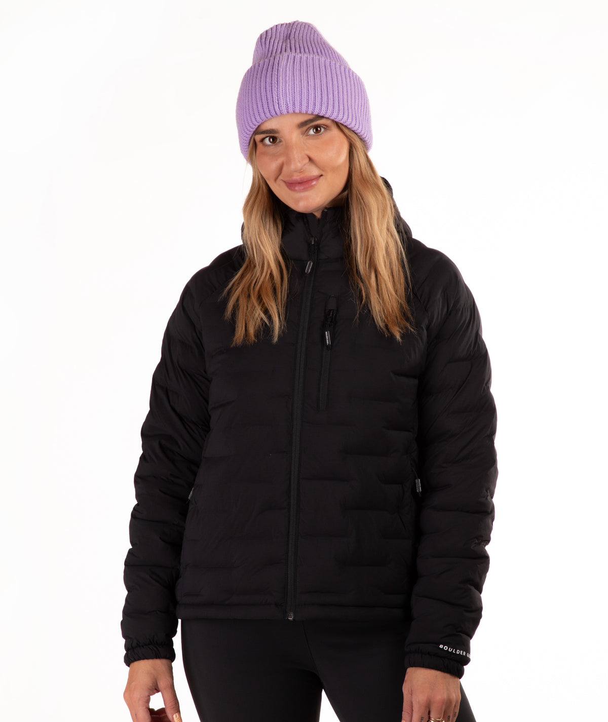 A woman wearing a black puffer jacket and a light purple ribbed knit beanie, standing against a plain white background.