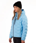 A woman wearing a light blue puffer jacket with black trim, a black beanie, and black pants, photographed against a plain white studio background.