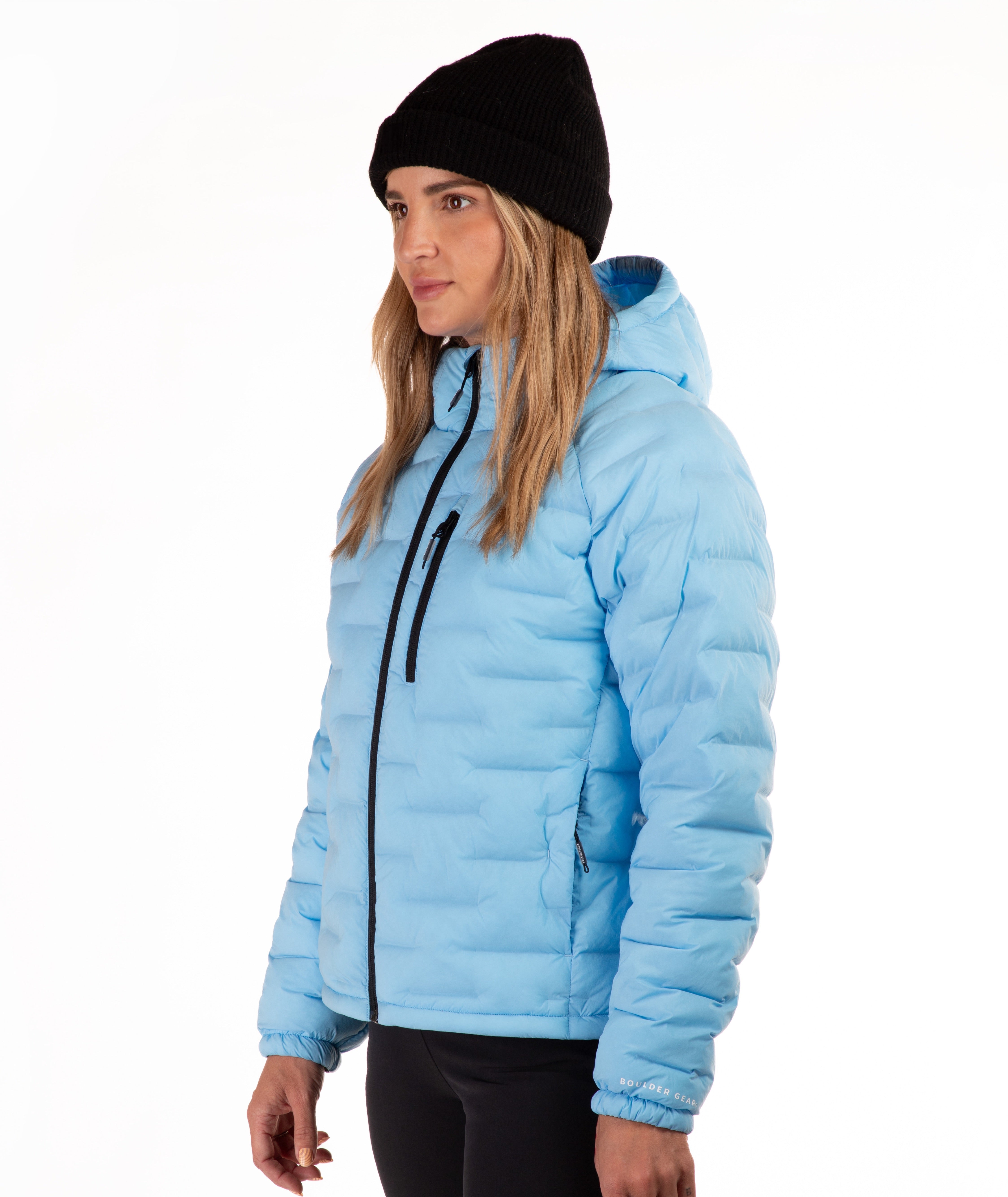 A woman wearing a light blue puffer jacket with black trim, a black beanie, and black pants, photographed against a plain white studio background.