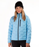 A woman wearing a light blue puffer jacket with black trim, a black beanie, and black pants, photographed against a plain white studio background.