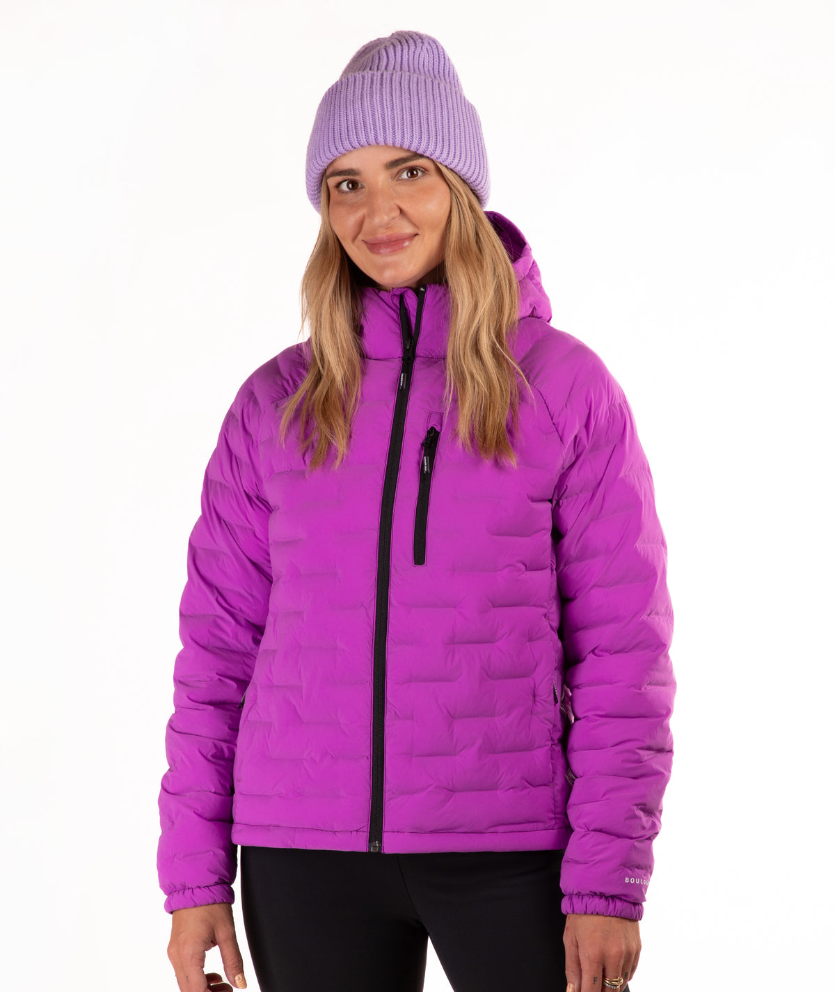 A woman wearing a bright purple quilted puffer jacket with a black zipper, a matching purple ribbed knit beanie, and black pants, photographed against a plain white studio background.