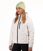 A woman wearing a off-white quilted puffer jacket with a black front zipper, paired with a light green ribbed knit beanie and black pants, photographed against a plain white studio background.