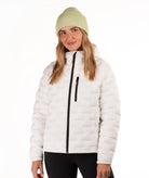 A woman wearing a off-white quilted puffer jacket with a black front zipper, paired with a light green ribbed knit beanie and black pants, photographed against a plain white studio background.