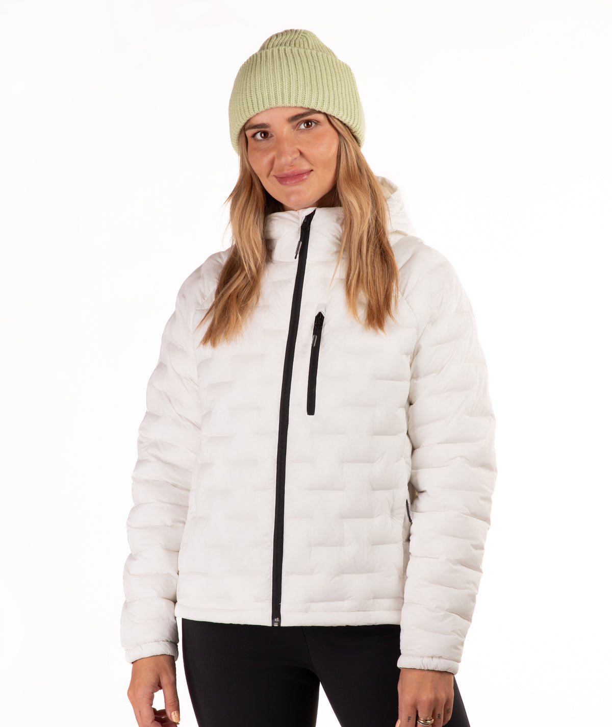 A woman wearing a off-white quilted puffer jacket with a black front zipper, paired with a light green ribbed knit beanie and black pants, photographed against a plain white studio background.
