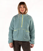 A woman wearing a light blue-green sherpa fleece pullover jacket with a yellow zipper, paired with dark brown pants, against a plain white background.