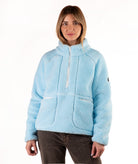 A woman wearing a light blue sherpa fleece pullover jacket with a half-zip neckline and front patch pockets, paired with dark brown pants, against a plain white background.