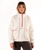 A woman wearing a off-white sherpa fleece pullover with red contrast stitching and a red half-zip, paired with dark brown jeans, against a plain white background.