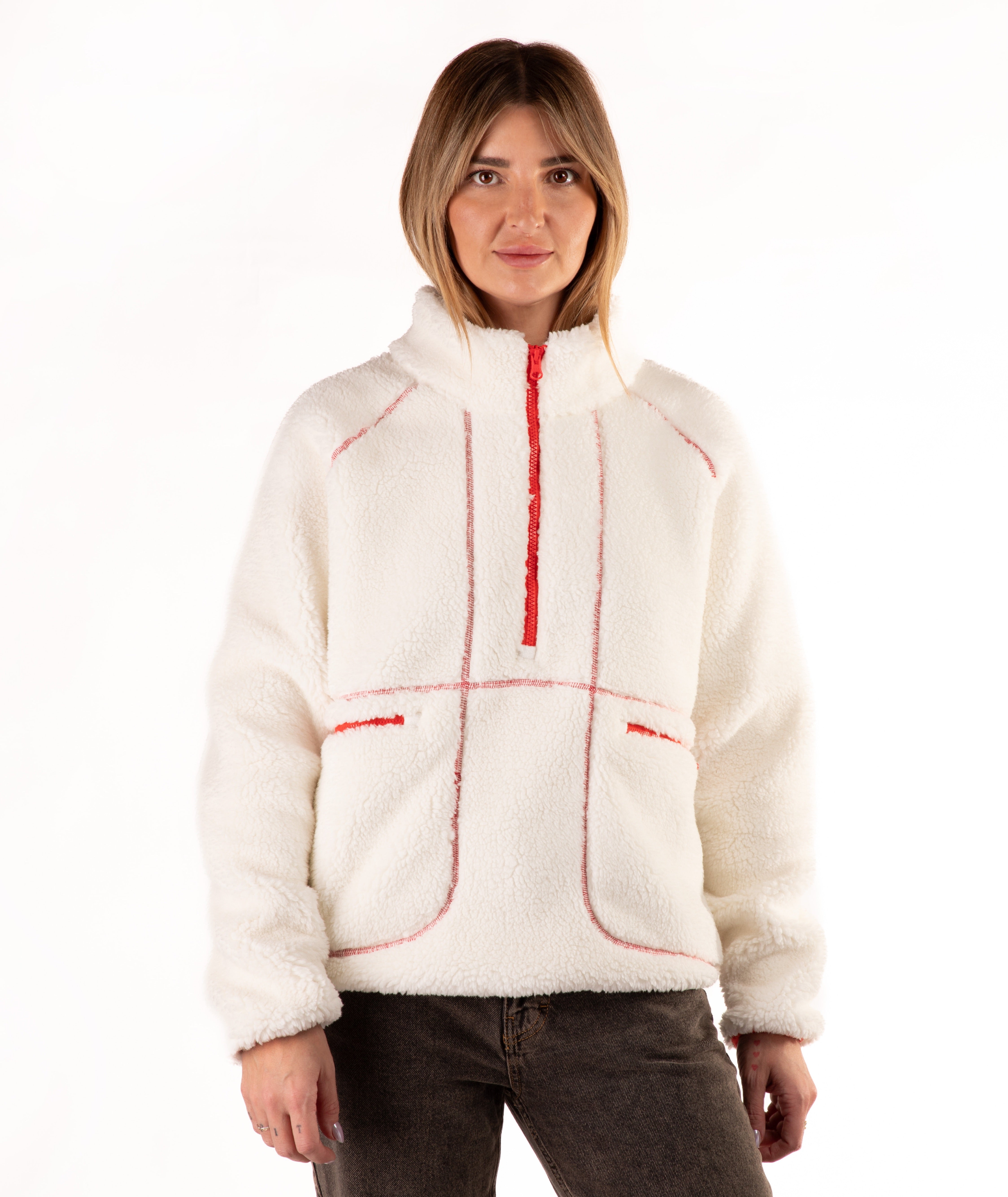 A woman wearing a off-white sherpa fleece pullover with red contrast stitching and a red half-zip, paired with dark brown jeans, against a plain white background.