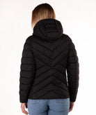 A woman viewed from the back wearing a black hooded puffer jacket with a distinctive chevron quilted pattern, paired with blue jeans, against a plain white background.