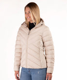 A woman wearing a light beige hooded puffer jacket with diagonal quilting, paired with blue jeans, photographed against a plain white background.