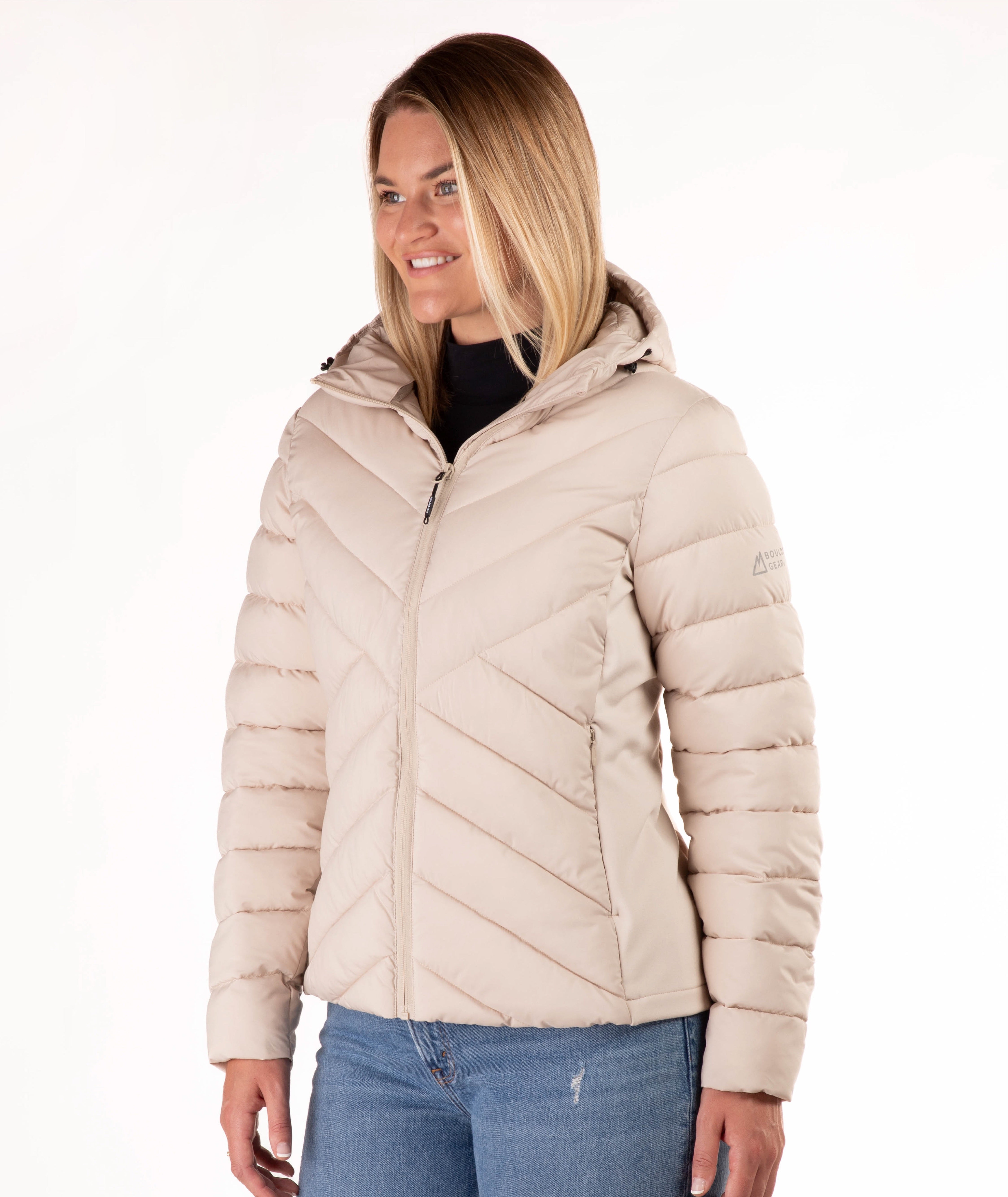 A woman wearing a light beige hooded puffer jacket with diagonal quilting, paired with blue jeans, photographed against a plain white background.