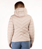 A woman viewed from the back wearing a light beige hooded puffer jacket with a chevron quilted pattern, paired with blue jeans, against a plain white background.