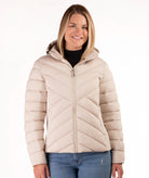 A woman wearing a light beige quilted puffer jacket with a hood, styled over a black top and blue jeans, against a plain white background.