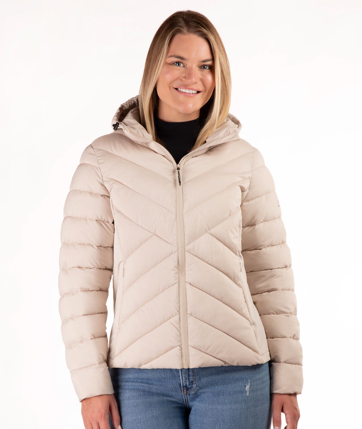 A woman wearing a light beige quilted puffer jacket with a hood, styled over a black top and blue jeans, against a plain white background.