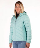 A woman wearing a light mint green hooded puffer jacket with diagonal quilting, paired with blue jeans, photographed against a plain white studio background.